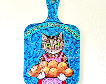 May include: A hand-painted wooden cutting board with a blue background and the words "PEACE LOVE CHALLAH". The board features a cat holding challah bread. The cat is wearing a pink apron. The Etsy shop name is visible.