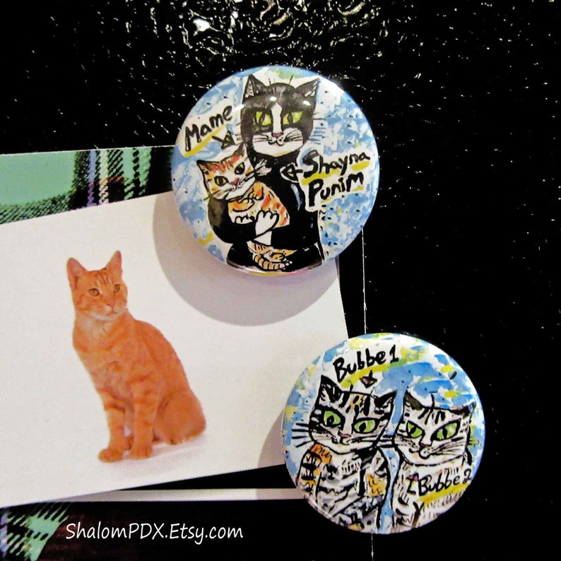 May include: Three round pinback buttons with colorful illustrations of cats. The first button features a black cat holding a smaller orange tabby cat with the text "Mame & Shayna Punim". The second button features two tabby cats with the text "Bubbe 1 & Bubbe 2".