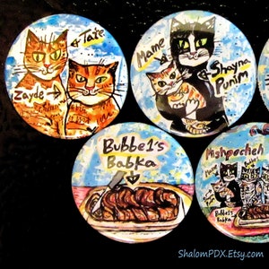 May include: Four colorful round buttons with painted images. The first button features two orange tabby cats with the names "Zayde" and "Tate". The second button features a black and white cat holding a smaller orange tabby cat with the names "Mame", "Shayna", and "Punim". The third button features a blue background with a brown babka and the text "Bubbe's Babka". The fourth button features a pink background with three cats and the text "Mishpoocheh".