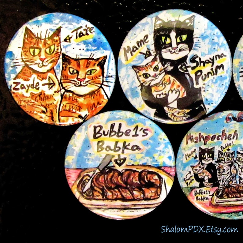 May include: Four colorful round buttons with painted images. The first button features two orange tabby cats with the names "Zayde" and "Tate". The second button features a black and white cat holding a smaller orange tabby cat with the names "Mame", "Shayna", and "Punim". The third button features a blue background with a brown babka and the text "Bubbe's Babka". The fourth button features a pink background with three cats and the text "Mishpoocheh".