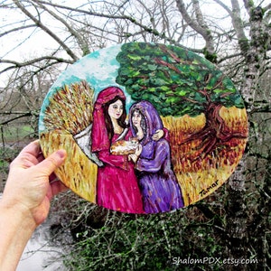 以下が含まれることがあります： 木製の丸い絵画で、カラフルなローブを着た2人の女性が小麦畑に立っている様子が描かれている。左側の女性は白い猫を抱いている。絵画は右下に「Tamar」と署名されている。