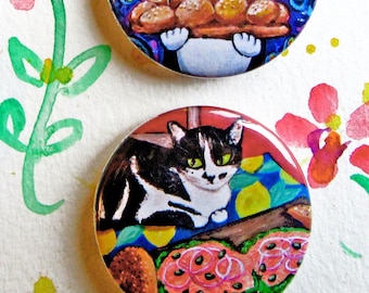 May include: Two round, colorful cat-themed buttons. One button features a black and white cat with green eyes, and the other shows a cat holding a tray of baked goods. The background includes watercolor flowers and the Etsy shop name.