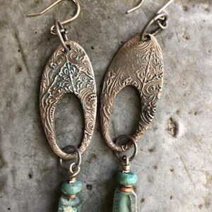 May include: A pair of oval-shaped earrings with an intricate etched design. The earrings are made of metal with a copper finish and have a turquoise bead dangling from each one.