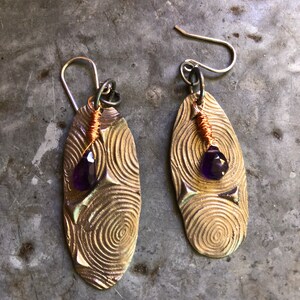 May include: A pair of silver earrings with a textured oval design and a purple gemstone in the center. The earrings have a wire wrapped around the top and a silver hook.
