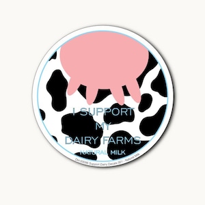 May include: A round sticker with a black and white cow print background. A pink cow udder is visible at the top of the sticker. The text "I SUPPORT MY DAIRY FARMS NATURAL MILK" is printed in blue on the sticker.