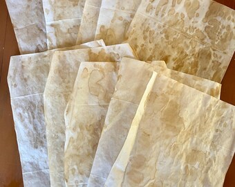 May include: A collection of folded, rectangular paper sheets with a vintage, aged appearance. The paper has a light brown color with darker brown stains and markings, giving it a rustic, antique aesthetic. The sheets are arranged in a layered pattern.
