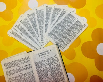 May include: An open English-French dictionary next to a fan of dictionary pages, all on a yellow floral background. The dictionary is open to a page of text. The fan of pages are curved and fanned out. The text "ENGLISH-FRENCH DICTIONARY" is visible.