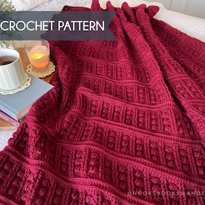 May include: A close-up of a red crocheted blanket with a textured pattern. The blanket is folded and draped over a bed with a book and a mug of tea on a wooden surface. The text "PDF CROCHET PATTERN" is visible at the top of the image.