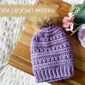 Könnte beinhalten: Lilafarbene Häkelmütze mit einem Kunstfellbommel. Die Mütze liegt auf einer Holzoberfläche mit einer rosa und lila Blume.