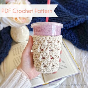 Pode incluir: Um protetor de copo de crochê bege sobre um fundo branco com o texto "Hooks, Books, & Wanderlust" na parte inferior. O protetor está em um copo de plástico transparente com um smoothie rosa dentro e um canudo vermelho.