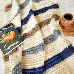 May include: A crocheted blanket with stripes of cream, brown, grey, and blue. The blanket is folded and draped over a white surface with a book and a mug on the side.