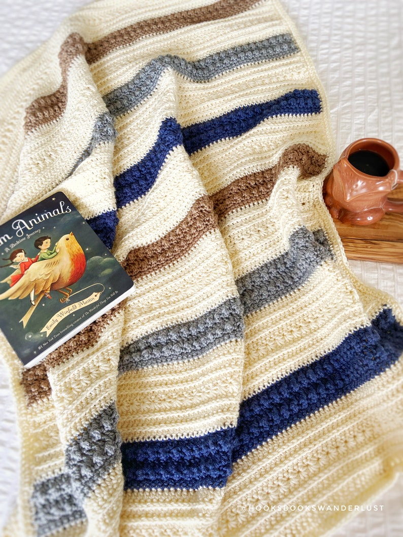 May include: A crocheted blanket with stripes of cream, brown, grey, and blue. The blanket is folded and draped over a white surface with a book and a mug on the side.