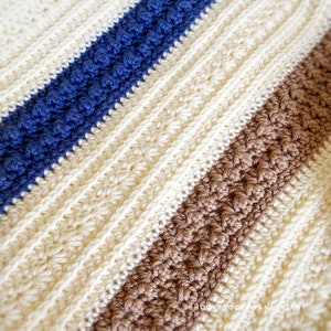 May include: A close-up of a crocheted blanket with stripes of cream, brown, and blue yarn. The blanket is made with a textured stitch that creates a raised pattern.