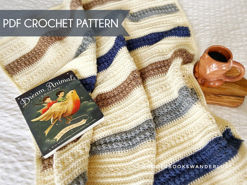 May include: A crocheted blanket with stripes of cream, brown, grey, and blue. The blanket is folded over a book titled "Dream Animals: A Bedtime Journey" by Emily Winfield Martin. A brown mug is sitting on a wooden surface.