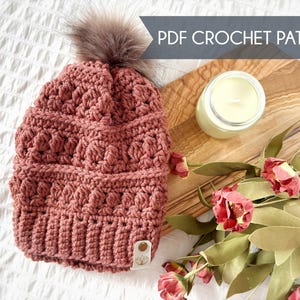 May include: A handmade, dusty rose crochet hat with a fluffy, brown pom-pom. The hat features a textured stitch pattern and a small tag. The image also includes a candle, flowers, and the text "PDF CROCHET PATTERN".