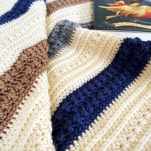 May include: A crocheted blanket with stripes of white, brown, blue and grey yarn. The blanket is folded and a book titled "Emily Winfield Martin" is on top of it.