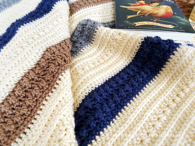 May include: A crocheted blanket with stripes of white, brown, blue and grey yarn. The blanket is folded and a book titled "Emily Winfield Martin" is on top of it.