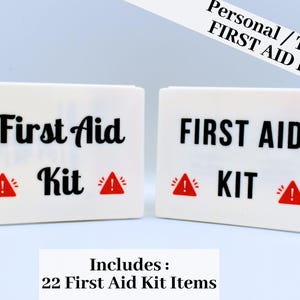 Puede incluir: Dos cajas blancas de botiquín con texto negro "First Aid Kit" y símbolos de peligro rojos. El texto "Personal / Travel FIRST AID KIT" está en la esquina superior derecha. El texto inferior dice "Includes: 22 First Aid Kit Items."