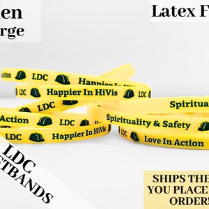 May include: Yellow rubber wristbands with black text and green construction worker helmet graphics. The text on the wristbands reads "LDC", "Happier In HiVis", "Spirituality & Safety", and "Love In Action".