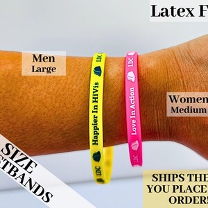 May include: Two silicone wristbands, one yellow with the text "Happier In HiVis" and the other pink with the text "Love In Action". The wristbands are on a person's arm. The text "Latex FREE" is at the top of the image. The text "SIZE WRISTBANDS" is at the bottom left of the image. The text "Men Large" is at the top left of the image. The text "Women Medium" is at the top right of the image. The text "SHIPS THE DAY YOU PLACE YOUR ORDER!!!!" is at the bottom right of the image.