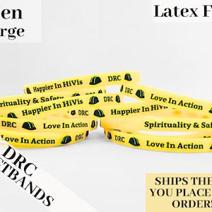 May include: A close-up of yellow silicone wristbands with black text and a green construction helmet graphic. The text on the wristbands reads "DRC", "Happier In HiVis", "Spirituality & Safety", and "Love In Action".