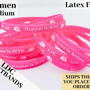 May include: A close-up of a stack of pink silicone wristbands with white text and a small white hard hat graphic. The text on the wristbands reads "LDC", "Happier In HiVis", and "Love In Action".
