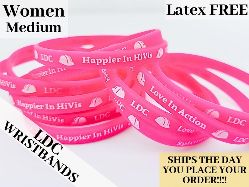 May include: A close-up of a stack of pink silicone wristbands with white text and a small white hard hat graphic. The text on the wristbands reads "LDC", "Happier In HiVis", and "Love In Action".