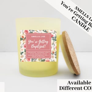 Puede incluir: Una vela amarilla en un tarro de vidrio con tapa de madera. La etiqueta dice "Smells Like You're Getting Baptized!" con detalles florales. La vela es de 85g.