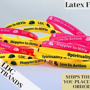 May include: A close-up of a pile of colorful silicone wristbands with various messages, including "Love In Action", "Happier In HiVis", "Spirituality & Safety", and "LDC".