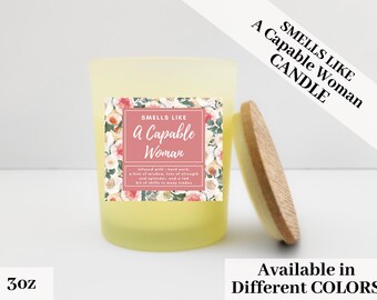May include: A yellow candle with a wooden lid. The candle has a label that reads "Smells Like a Capable Woman." The label is surrounded by a floral design. The candle is 3 oz.