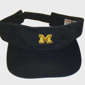 May include: Black visor with a gold embroidered "M" logo. The visor has a curved brim and an adjustable strap. The visor is made of a durable fabric and is designed to shield the eyes from the sun. The visor is a sports accessory.