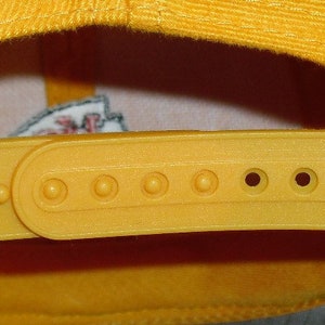 May include: A yellow baseball cap with a yellow adjustable strap and a row of holes for adjusting the size. The strap is made of a smooth, plastic-like material.