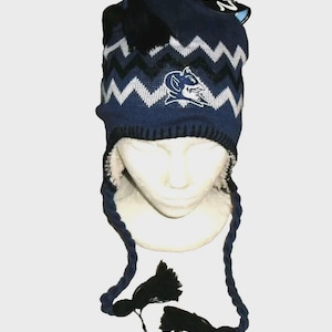 May include: A navy blue knit hat with a white and black zigzag pattern. The hat features a white embroidered logo and a fleece lining. It has braided ties with black tassels. A winter hat.
