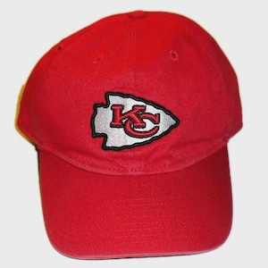 May include: A bright red baseball cap featuring an embroidered arrowhead-shaped patch on the front. The patch is white with a black border and contains the red letters KC in a stylised font. The cap has a curved brim and ventilation eyelets.