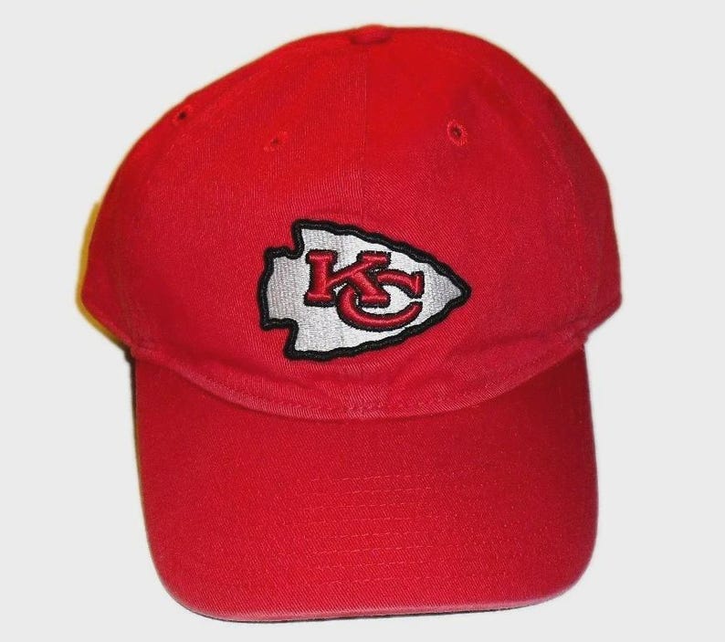 May include: A bright red baseball cap featuring an embroidered arrowhead-shaped patch on the front. The patch is white with a black border and contains the red letters KC in a stylised font. The cap has a curved brim and ventilation eyelets.