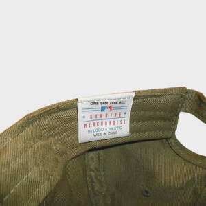 May include: An olive green baseball cap featuring an interior white fabric tag. The tag displays the text ONE SIZE FITS ALL, a small MLB logo, GENUINE MERCHANDISE, By LOGO ATHLETIC, and MADE IN CHINA, all printed in blue and red ink.