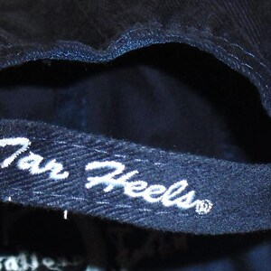 K&ouml;nnte beinhalten: Eine dunkelblaue Baseballkappe mit einem wei&szlig;en gestickten Logo, auf dem "Tar Heels" steht.