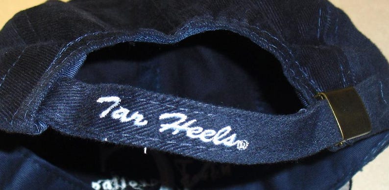 K&ouml;nnte beinhalten: Eine dunkelblaue Baseballkappe mit einem wei&szlig;en gestickten Logo, auf dem "Tar Heels" steht.