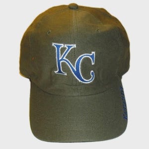 May include: An olive green baseball cap featuring the letters KC embroidered in blue with a white outline on the front panel. The word KANSASCITY is embroidered in blue vertically along the right side of the curved brim of the cap.