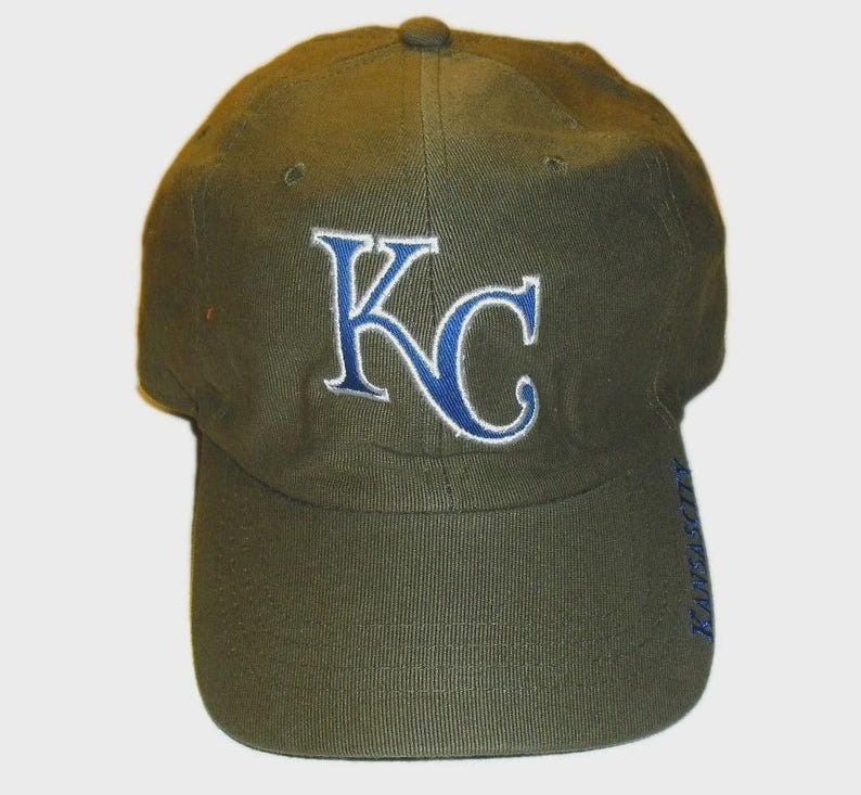 May include: An olive green baseball cap featuring the letters KC embroidered in blue with a white outline on the front panel. The word KANSASCITY is embroidered in blue vertically along the right side of the curved brim of the cap.