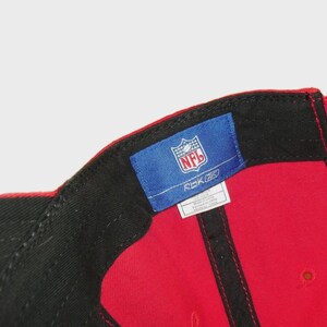 May include: A red baseball cap featuring a black interior sweatband and a blue rectangular tag sewn onto the inner crown. The tag displays the NFL logo in white and red, the Reebok logo, and a small white label with printed text details.