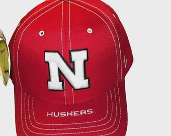 May include: A red baseball cap with a white "N" logo and the word "HUSKERS" embroidered on the front. The cap features white stitching and a curved brim. A classic sports fan accessory.