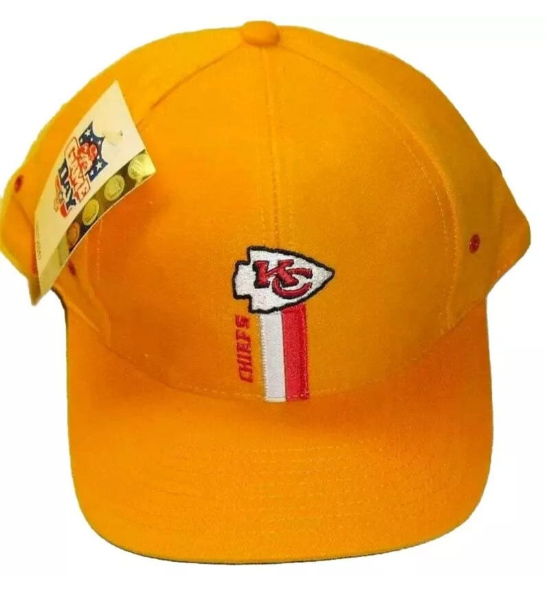 May include: A yellow baseball cap with the Kansas City Chiefs logo and the words "Chiefs" embroidered on the front. The cap has a red, white, and blue stripe down the center.
