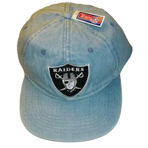 May include: A light blue denim baseball cap with a black and white Raiders logo. The logo features a skull with crossed swords and the word "RAIDERS" above it. The cap has a Team NFL tag attached to the side.