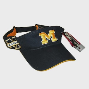 May include: A navy blue visor featuring a yellow embroidered block M on the front and the word MICHIGAN embroidered on the side. The visor has a yellow trim along the brim and includes a rectangular product tag attached to the side strap.