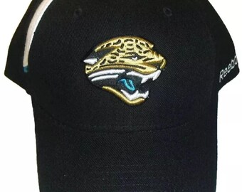 May include: Black baseball cap with a white stripe and a gold embroidered Jacksonville Jaguars logo. The logo features a jaguar's head with a teal and white background. The text "Reebok" is visible on the right side of the cap.