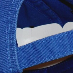 May include: Royal blue baseball cap with the embroidered letters "KC" in gold. The cap has a curved brim and an adjustable strap with a brass buckle. The underside of the brim is white.