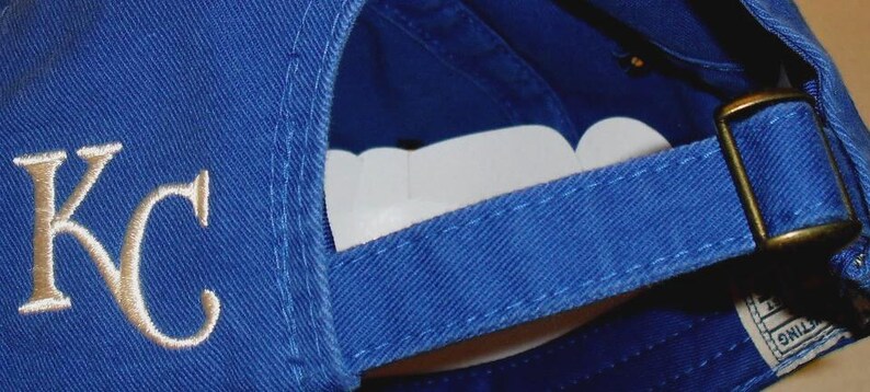May include: Royal blue baseball cap with the embroidered letters "KC" in gold. The cap has a curved brim and an adjustable strap with a brass buckle. The underside of the brim is white.