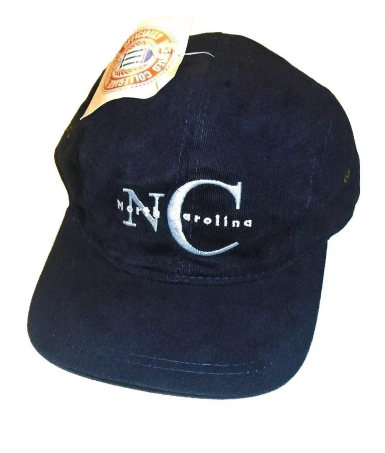 K&ouml;nnte beinhalten: Eine marineblaue Baseballkappe mit einem wei&szlig;en gestickten "NC"-Logo und dem Wort "Carolina" darunter. Die Kappe hat einen gebogenen Schirm und ein wei&szlig;es Etikett, das an der Seite befestigt ist.