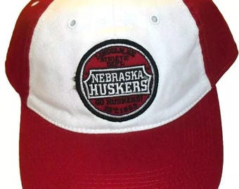 May include: A red and white baseball cap with the text "Nebraska Huskers" on the front. The cap has a curved brim and a structured crown. The cap has a tag attached to the top.